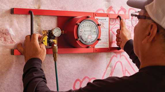 A close-up shot of an insulation technician using a specialized gauge and tools to measure pressure and airflow in a home's interior wall. The image highlights the detailed quality assurance and inspection process that ensures the insulation job is done correctly.